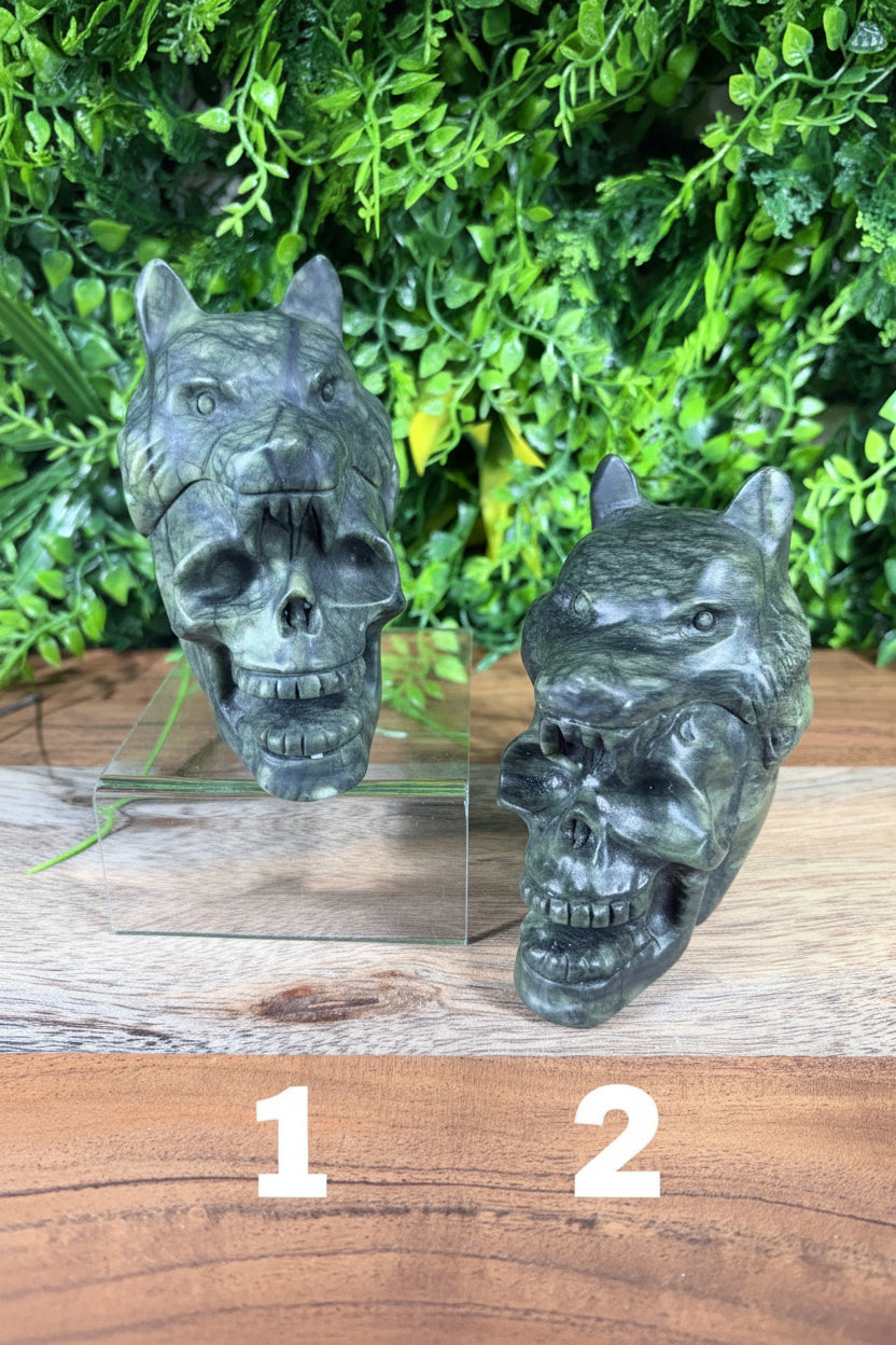 Two dark green stone wolf skull sculptures displayed on a clear acrylic stand against a background of green foliage and a wooden surface.