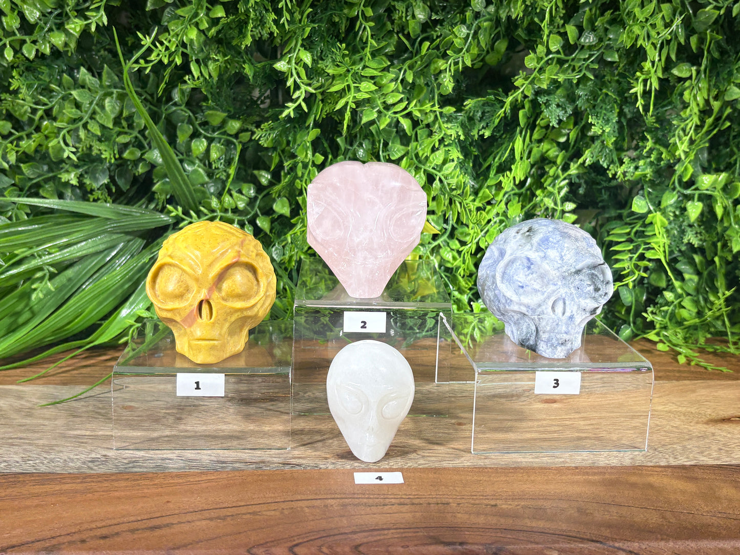 Four carved crystal stone figurines arranged on clear acrylic display stands against a green foliage backdrop. The set includes a yellow skull, a pink heart, a blue-gray skull, and a white alien head, each labeled with a small number plaque.