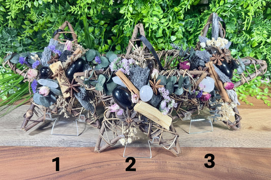 Three decorative star-shaped crystal clusters made from woven branches, black and polished white stones, dried lavender, cinnamon sticks, and small dried flowers, displayed on clear stands against a green foliage backdrop and wooden surface.