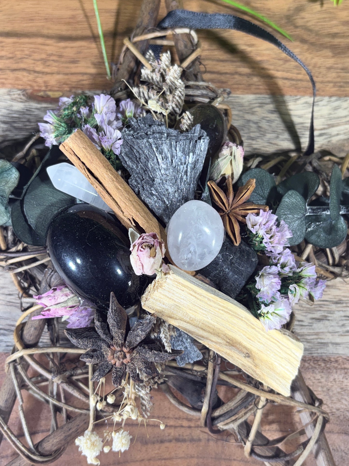 A dried botanical and crystal arrangement featuring a polished black obsidian, a smooth white quartz crystal, a gray mineral specimen, cinnamon sticks, star anise, small purple flowers, and natural wood pieces on a rustic wood background.