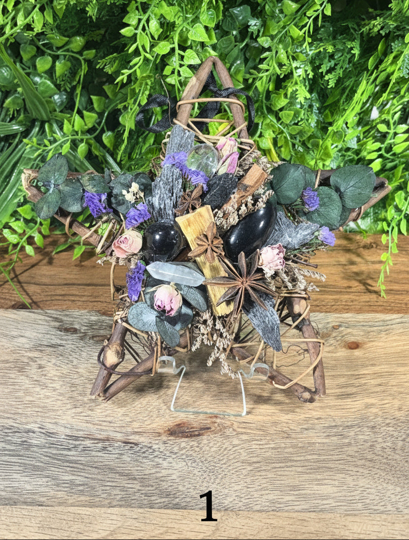 A rustic dried floral arrangement on a small metal stand, featuring dark brown twigs, green leaves, purple and pink flowers, star anise, cinnamon sticks, and black stones, set against a green leafy background and wood surface.