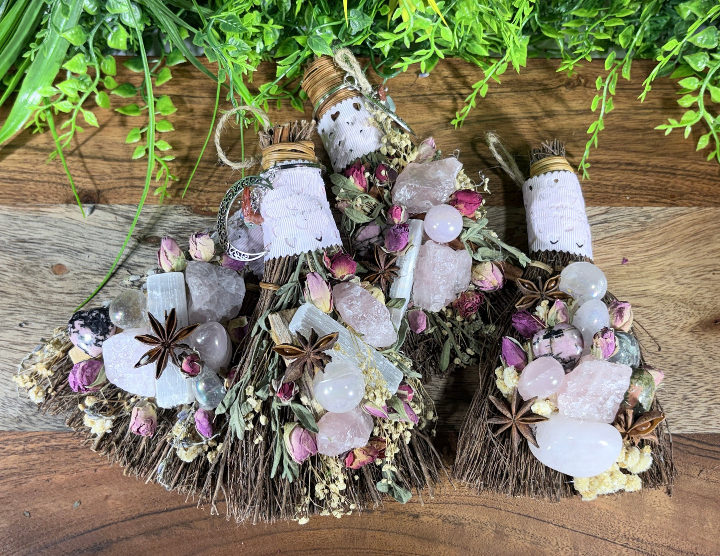 Two decorative broomsticks made from dried grasses, wrapped in white paper, and adorned with crystals, rosebuds, star anise, and herbs, displayed on a wooden surface with green foliage in the background.