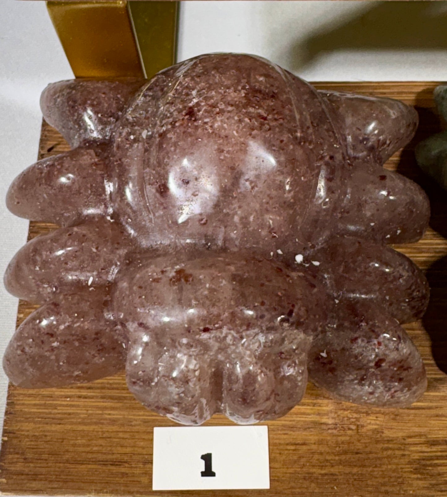 A carved translucent pinkish-brown stone sculpture with a rounded organic shape and textured speckles, displayed on a wooden surface with a small white label marked '1'.