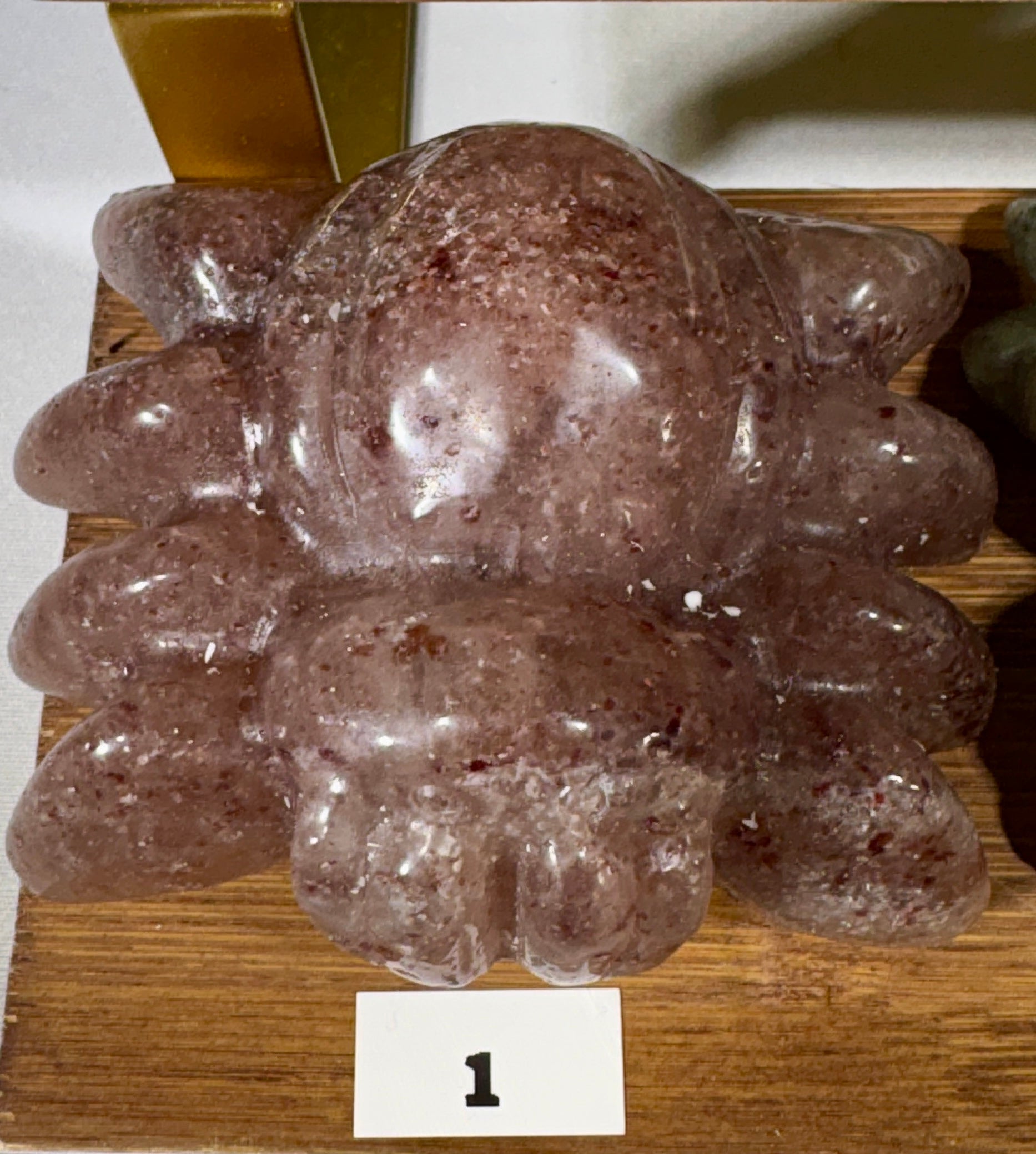 A carved translucent pinkish-brown stone sculpture with a rounded organic shape and textured speckles, displayed on a wooden surface with a small white label marked '1'.