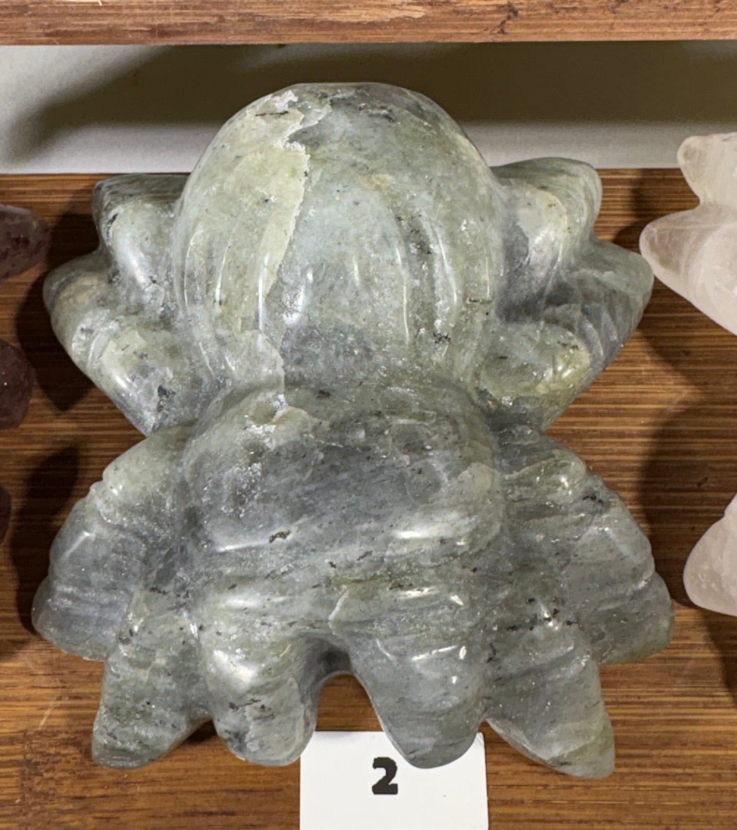 A carved translucent gray stone sculpture with a rounded organic shape, displayed on a wooden surface with a small white card marked '2' below it.