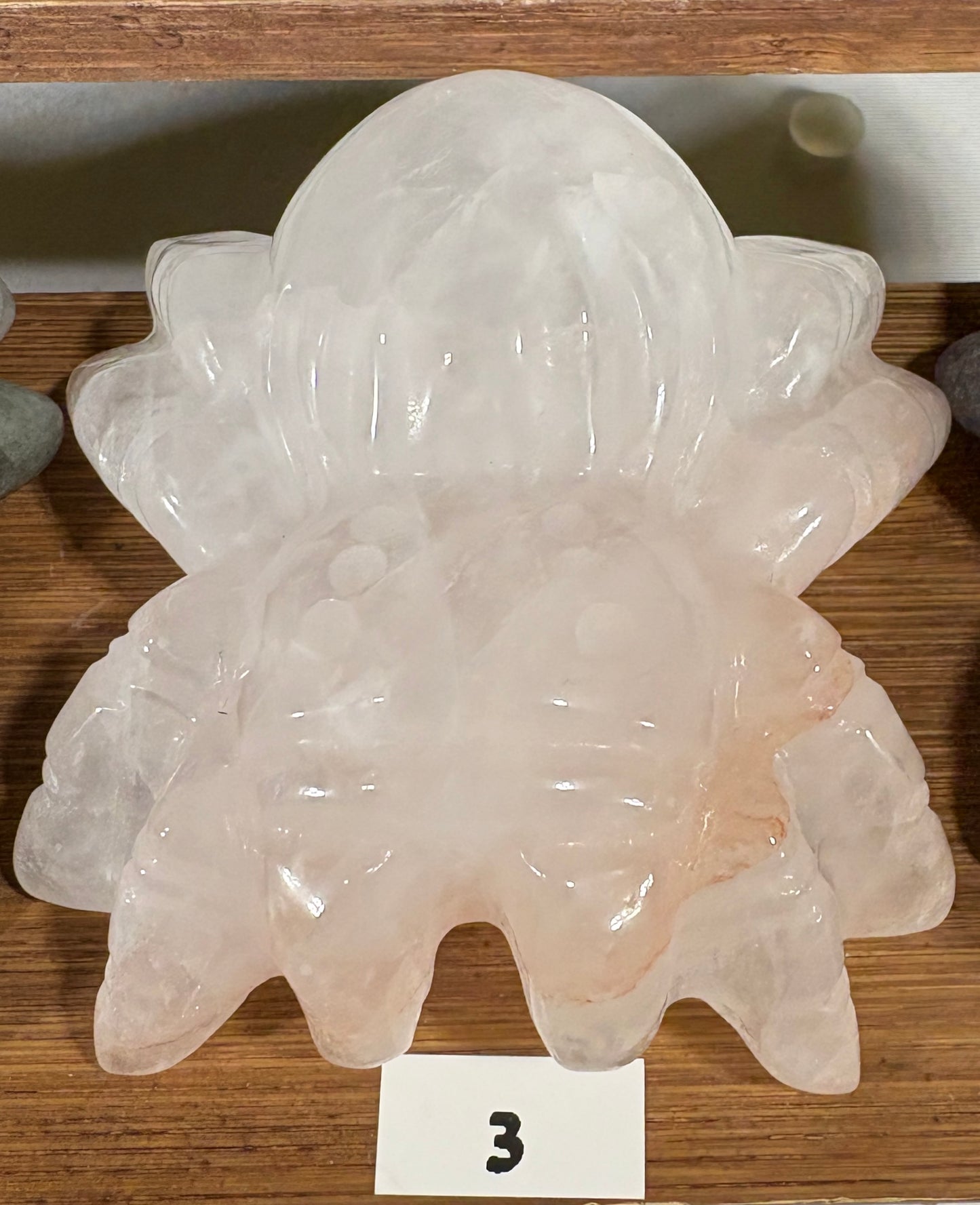 A carved pale pink stone figurine with a rounded, organic shape and textured surface, displayed on a wooden surface with a small white card marked '3' in front.