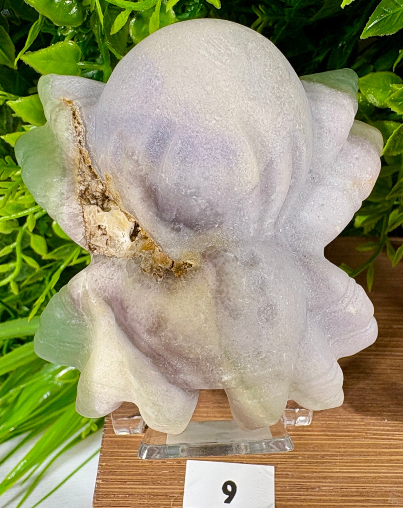 A carved pale lavender and off-white stone spider figurine displayed on a small clear stand, with green foliage and a wood surface in the background.