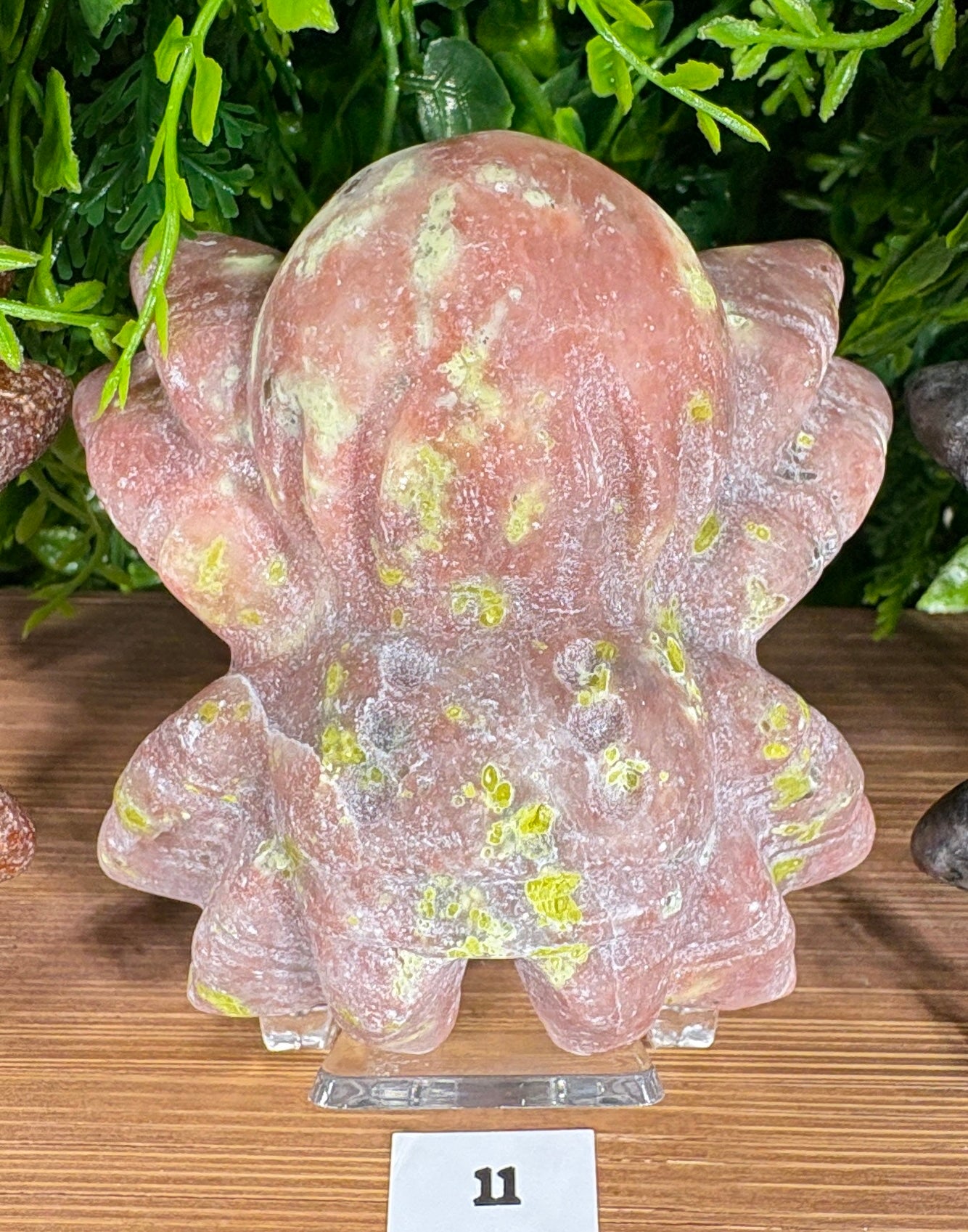 A carved pink stone spider figurine with yellow-green marbled inclusions, displayed on a wooden surface with green foliage in the background.