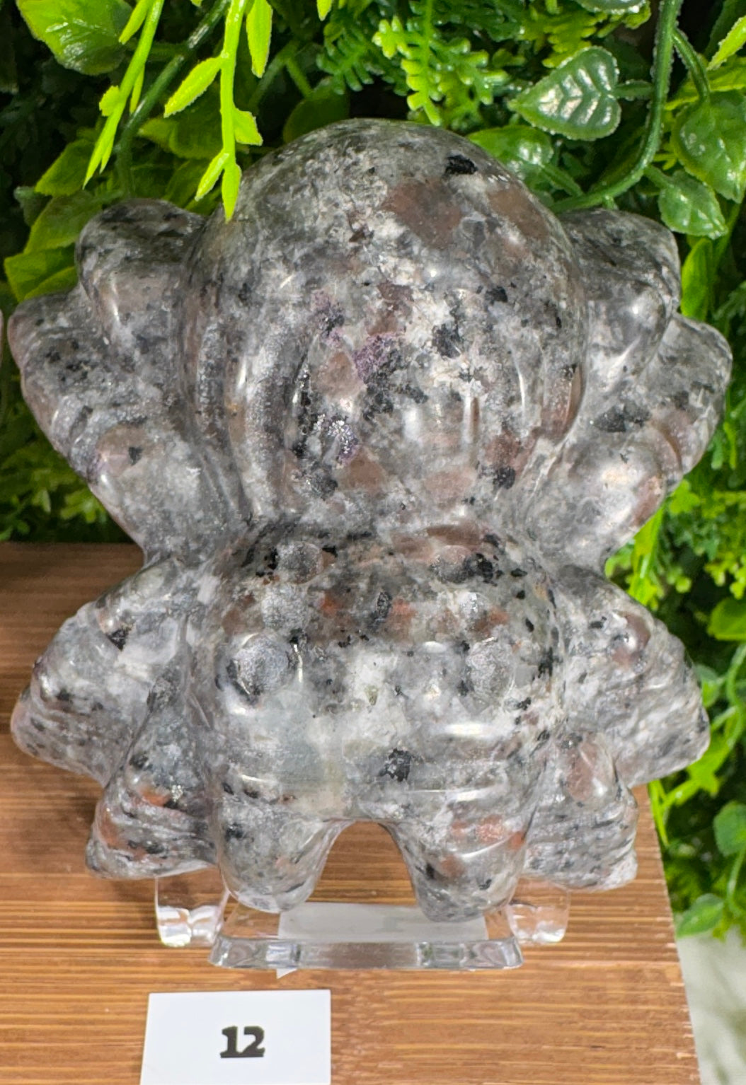 A carved crystal spider with a textured, polished surface in gray, black, and reddish-brown tones, displayed on a clear acrylic stand on a wooden surface with green foliage in the background.