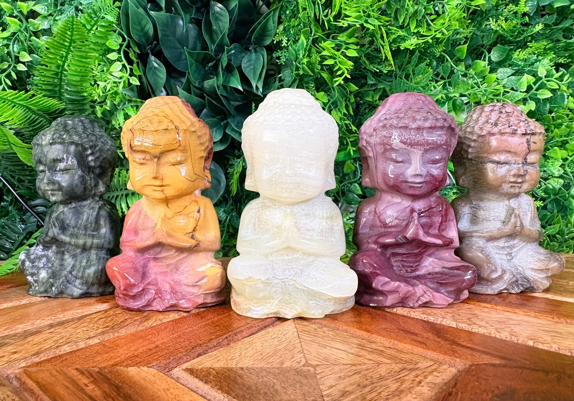 Five colorful stone statues of Buddha on a wooden surface with green foliage in the background