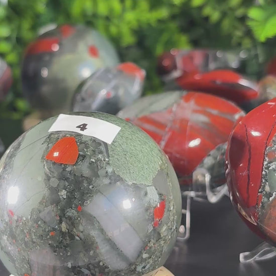 Video of  polished African bloodstone crystal spheres, palm stones and pointed crystal towers on a rotating stand , set against a dense green foliage backdrop. 