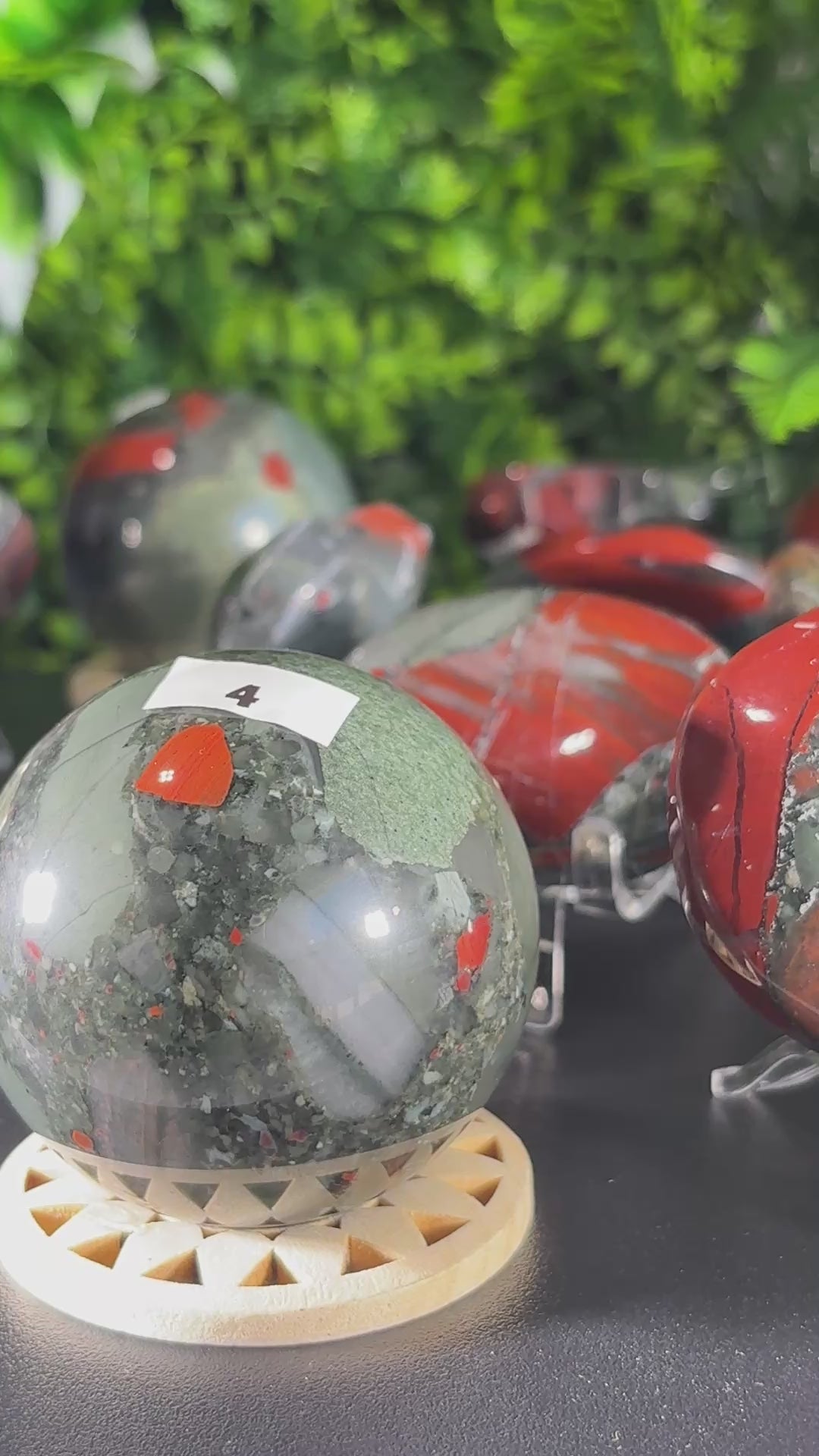 Video of  polished African bloodstone crystal spheres, palm stones and pointed crystal towers on a rotating stand , set against a dense green foliage backdrop. 